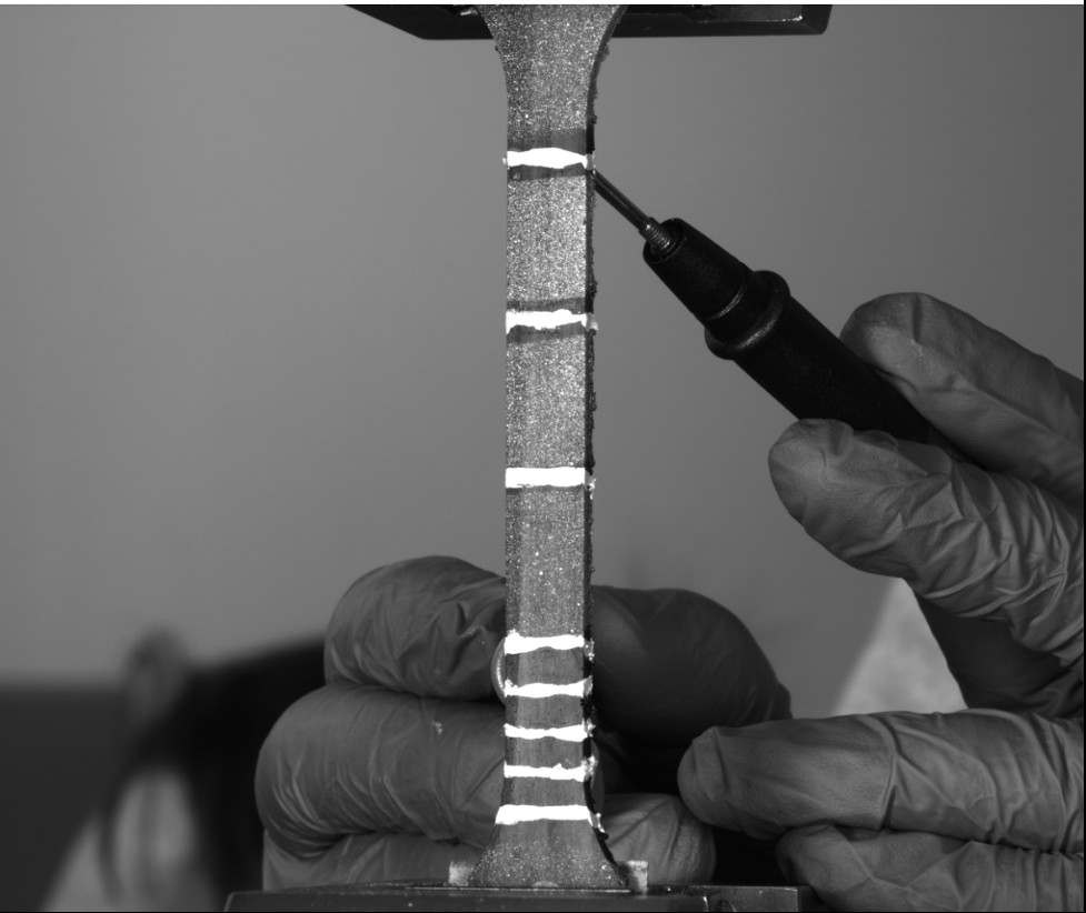 CNT-nanocomposites being tested for piezoresistive response in an Intron 5969 Universal Testing Systems under tensile load. The values of resistance change as increasing tensile load is applied which allow the material to send a self-report as to its condition. The silver lines are conductive contacts and the white speckles are used by the LIMESS digital image correlation (DIC) system to calculate strain values. Image was taken in the Laboratory of Mechanical Testing, CDMM.
